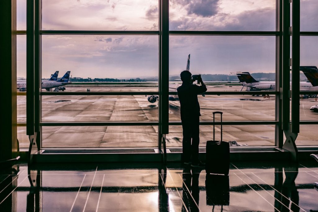 Traveler using a phone to plan a trip with AI, silhouette against airport window watching a plane