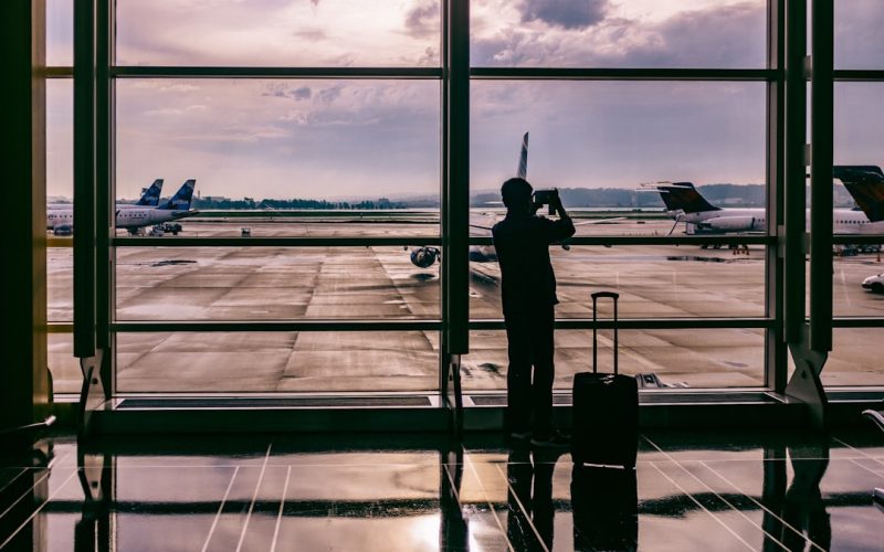Traveler using a phone to plan a trip with AI, silhouette against airport window watching a plane